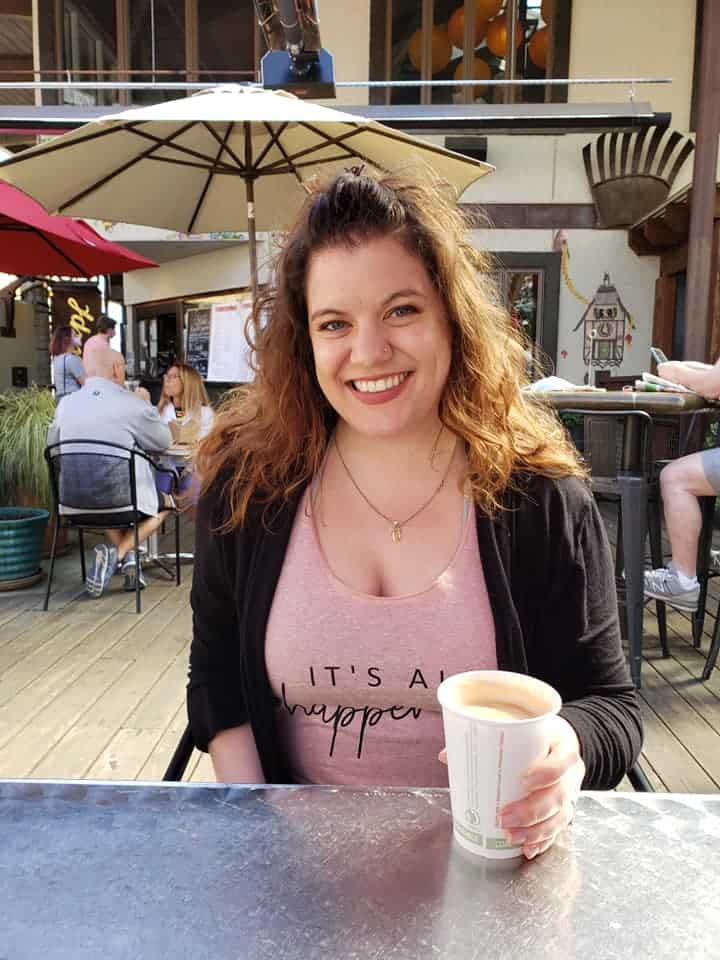 Amy Hartle enjoys a leisurely moment at an outdoor café in North Carolina, her radiant smile matching the sunny ambiance, as she holds a warm cup of coffee, epitomizing a relaxed lifestyle 