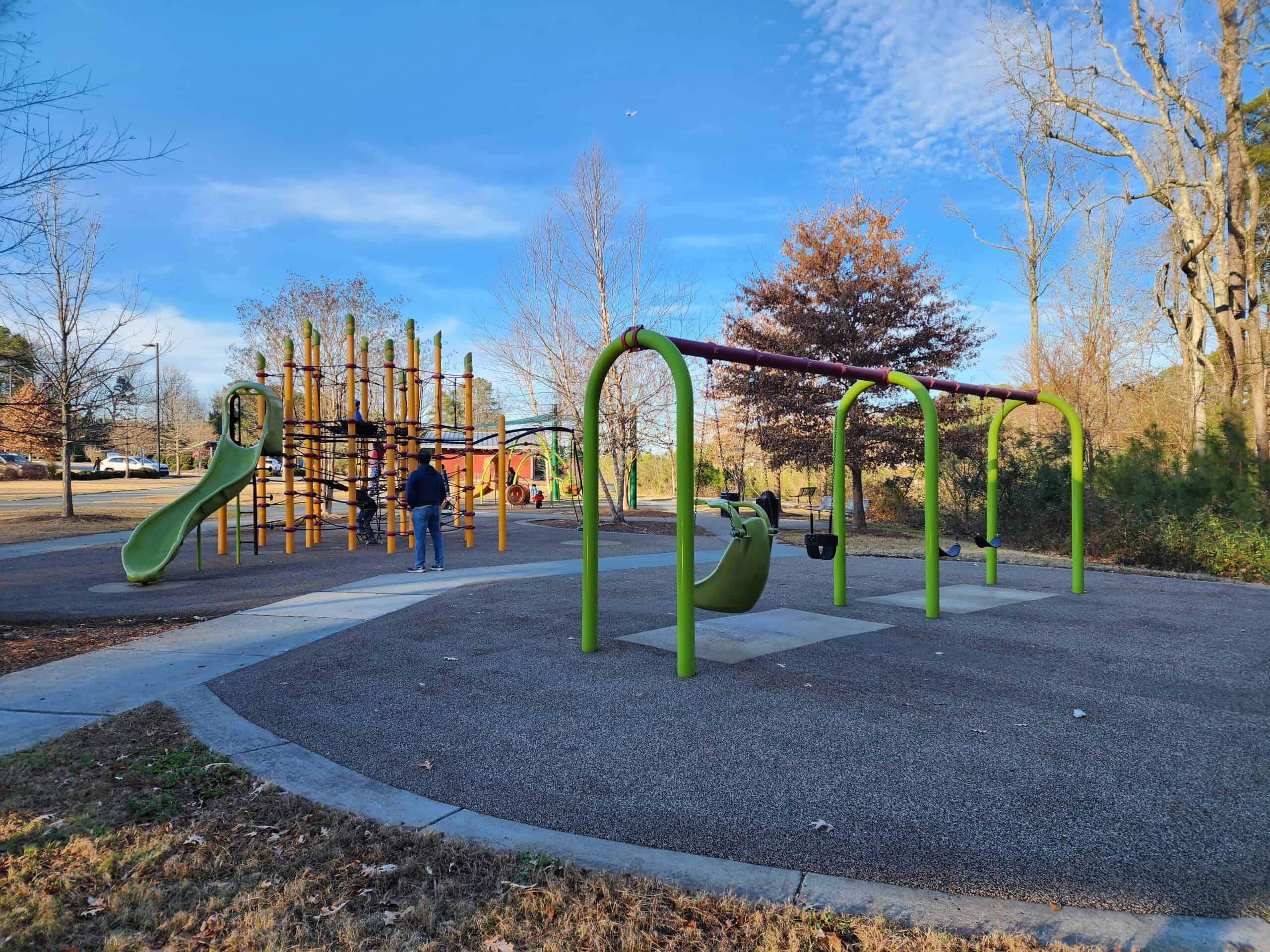 A vibrant playground at Carpenter Park in Cary, North Carolina, with colorful play structures including swings and a slide, set against a backdrop of bare winter trees under a clear blue sky, showcasing the park's recreational amenities for a playground review