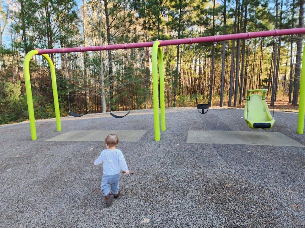 A young child approaches the swing set at Carpenter Park in Cary, with the playground's slide in the background, surrounded by tall pine trees, illustrating a family-friendly and inviting outdoor play area.
