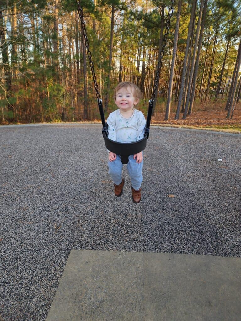 A delighted toddler in a swing at Carpenter Park in Cary, smiling joyfully, with the peaceful backdrop of a pine forest highlighting the park's serene and enjoyable environment for children.