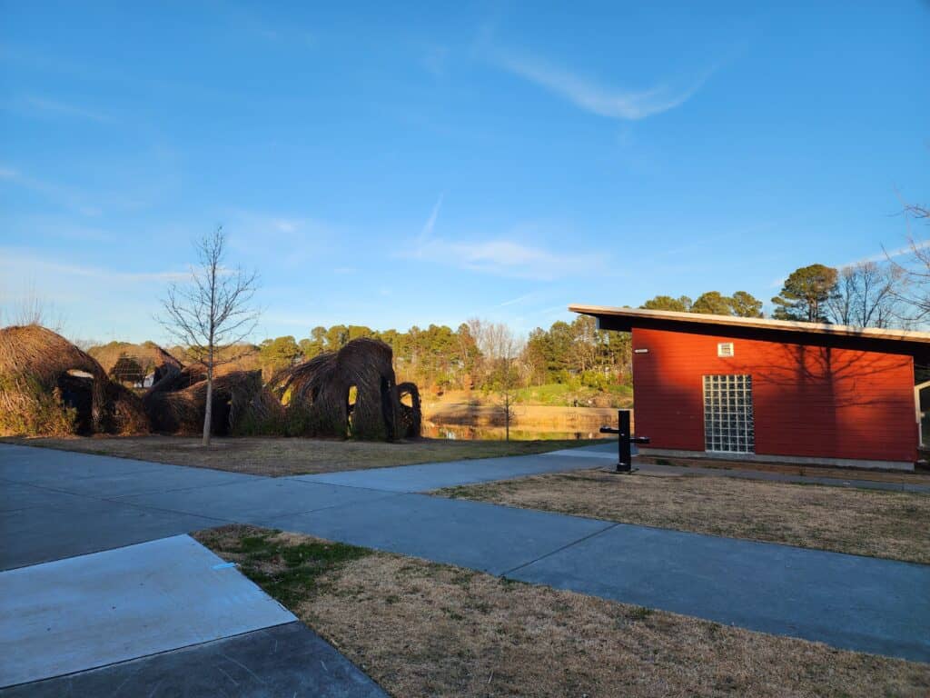 Late afternoon sun casts a warm glow on a public restroom facility near a unique playground with natural woven sculptures at a park in Cary, North Carolina, reflecting a calm and inviting community space.