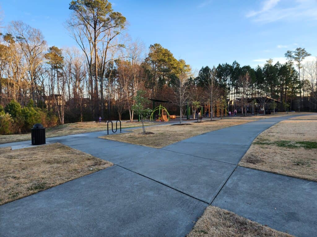 Late afternoon at Carpenter Park in Cary, North Carolina, featuring a quiet playground with a walking path, bicycle racks, and surrounding tall trees that signal the approach of spring, ideal for a detailed playground review