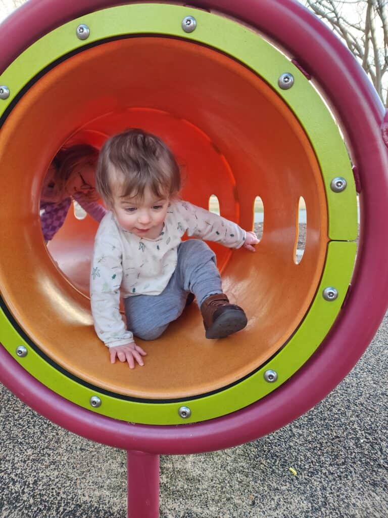 An adorable moment captured as a toddler crawls through a colorful tunnel at Carpenter Park in Cary, showcasing the engaging and playful equipment available for young children at this local playground