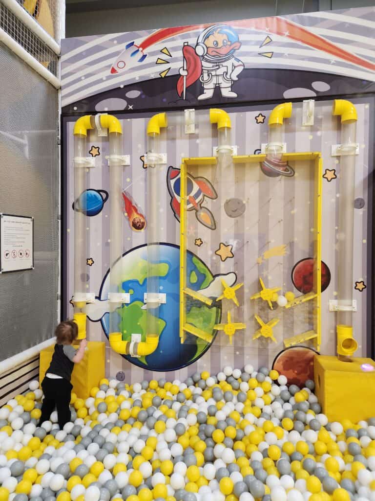 A toddler interacts with a colorful space-themed wall game at an indoor playground in the Triangle, surrounded by a sea of yellow and white balls