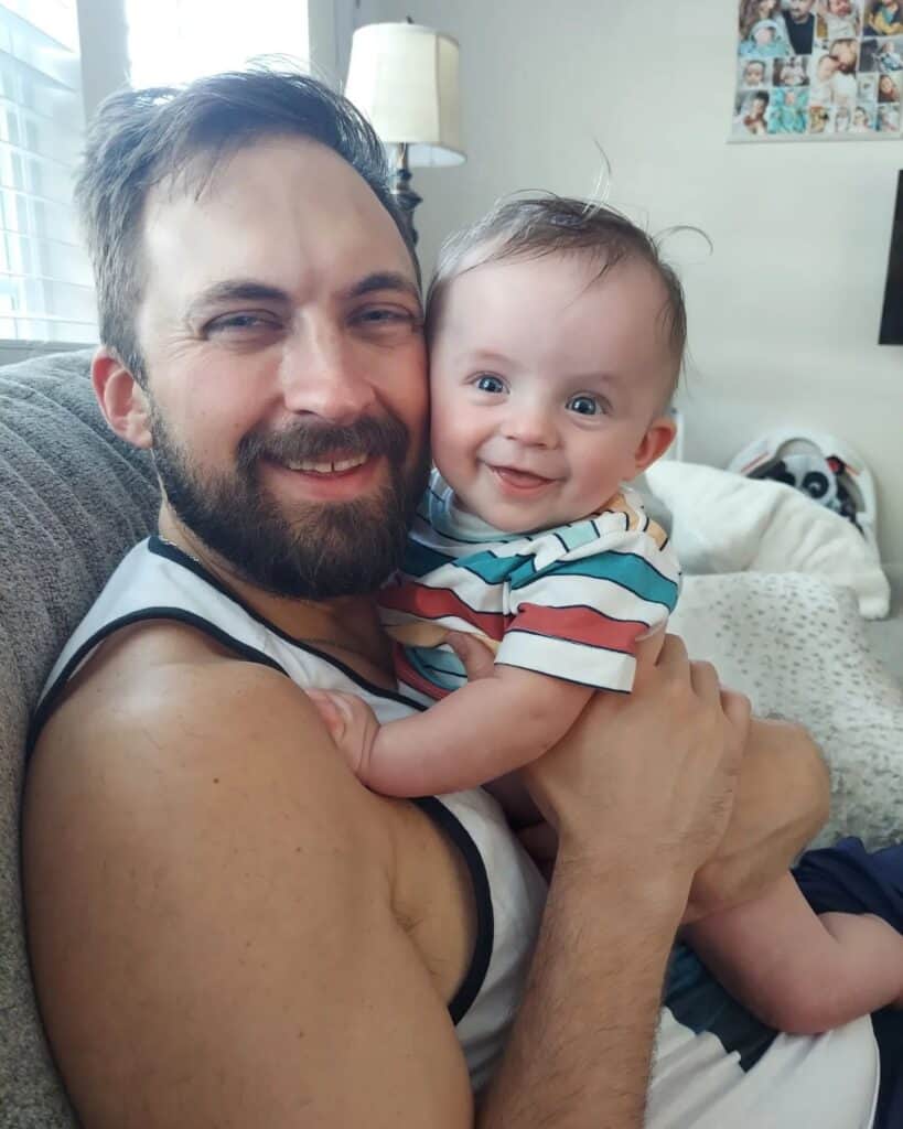 Nathan Hartle shares a heartwarming embrace with his infant, both smiling joyfully, capturing a serene father-child moment at home, with the baby's bright eyes and playful grin highlighting the bond they share