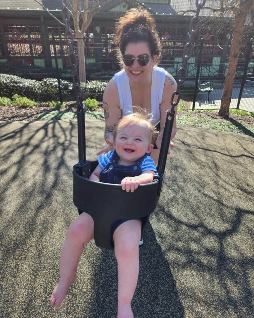 An ecstatic toddler with a beaming smile enjoys a swing ride, gently pushed by a smiling woman with sunglasses, at Pullen Park in Raleigh, NC, capturing a moment of pure joy and family fun in this beloved local park.