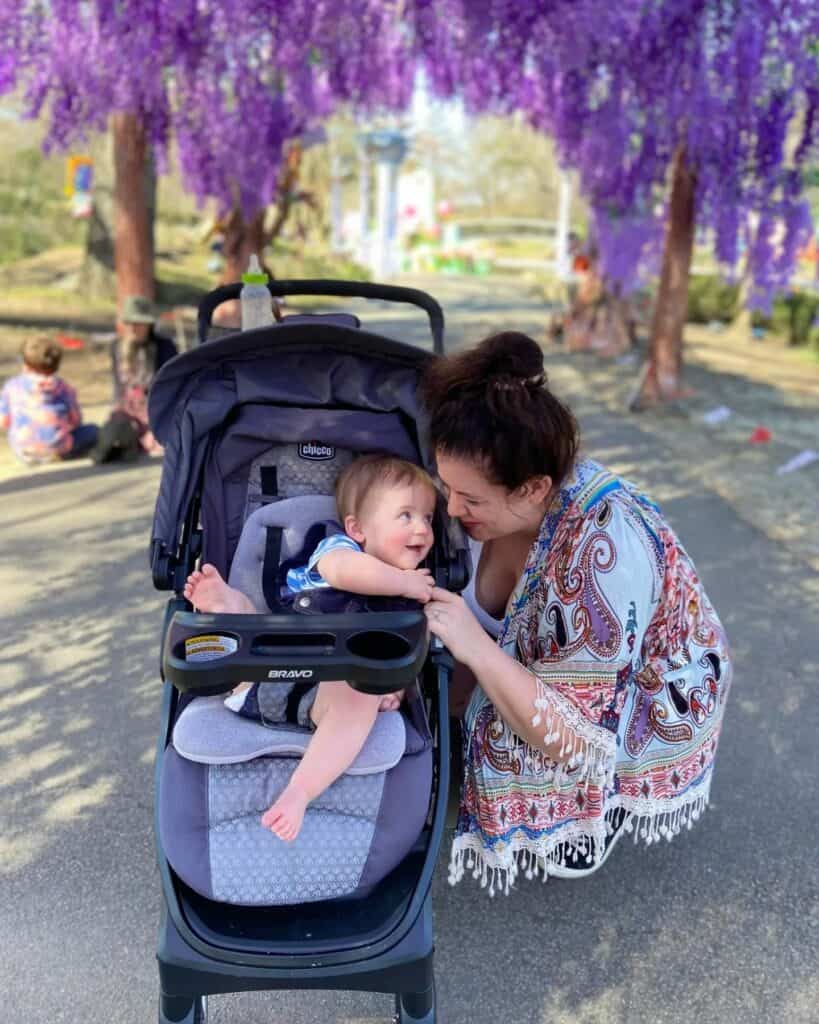 In the midst of a North Carolina family outing, a woman in a patterned shawl shares a tender moment with her baby, who sits in a stroller, both enchanted by each other under the whimsical purple blooms of a wisteria tree.