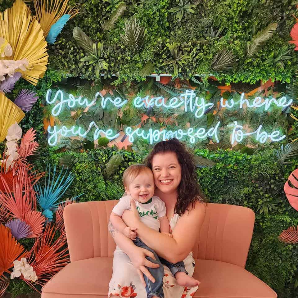 A joyful moment captured in Raleigh, with a smiling woman in a floral dress holding a giggling toddler, both seated against a vibrant backdrop of lush greenery and neon sign saying 'you're exactly where you're supposed to be', embodying the spirit of a Raleigh family adventure