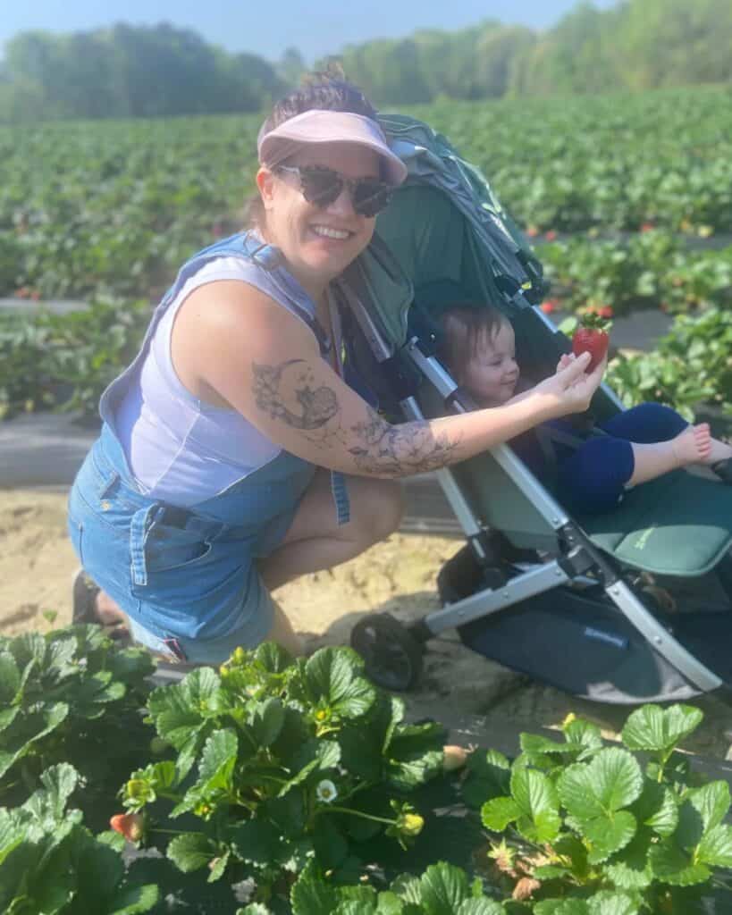 A radiant mother in a denim jumpsuit and sunglasses kneels beside a stroller, offering a large ripe strawberry to her delighted child, amidst a vast strawberry field, highlighting family-friendly activities in the Triangle area