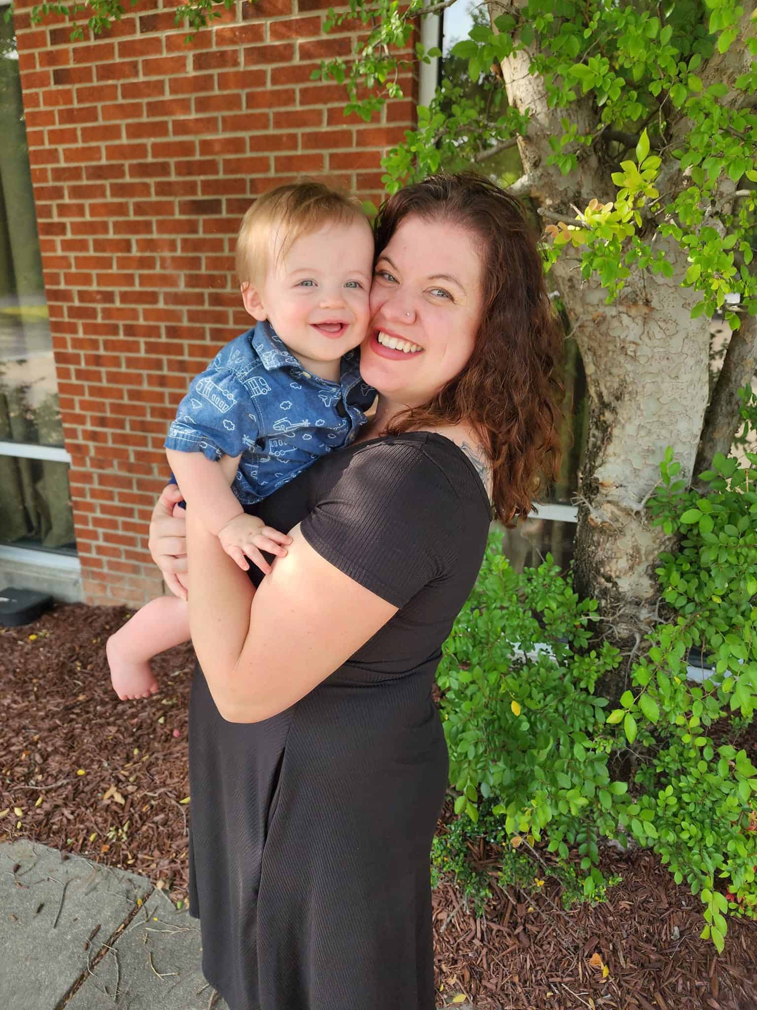 A Raleigh mom blogger shares a sunny embrace with her cheerful toddler, both radiating happiness in a casual outdoor setting with green foliage and a brick backdrop, epitomizing the warmth and joy of family life in Raleigh