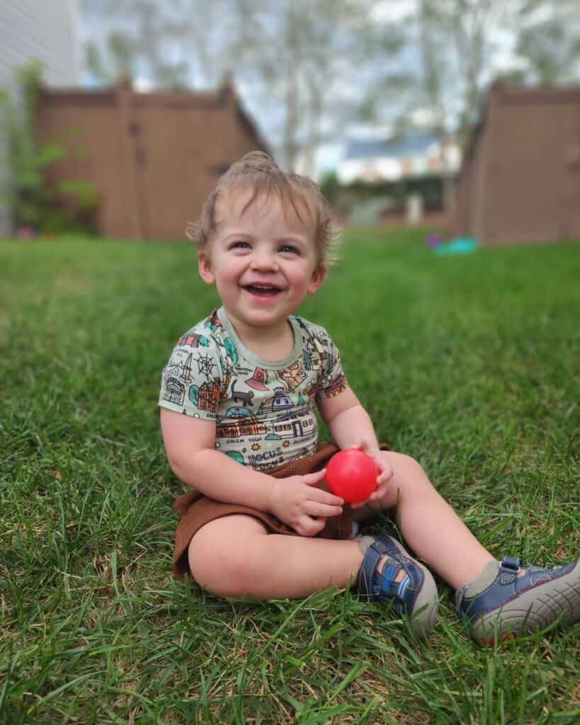 Malcolm, a joyful toddler with tousled hair, sits on the grass clutching a red ball, his face lit up with a delightful smile, capturing a moment of pure childhood bliss and playful discovery outdoors."


