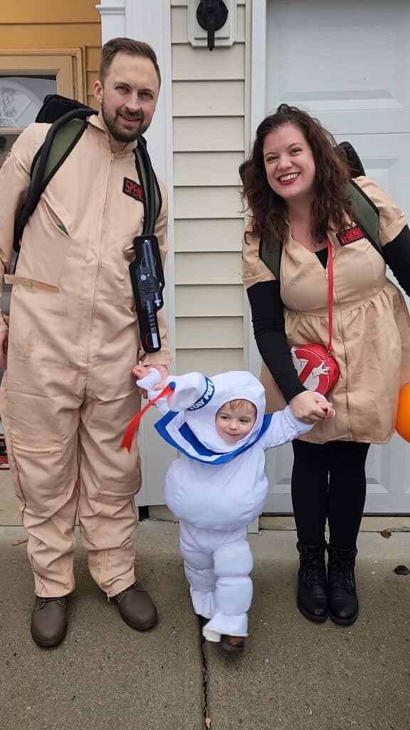 The Hartle family steps out in themed costumes, with the parents dressed as ghostbusters and their toddler adorably costumed as a mini Stay Puft Marshmallow Man, ready for a playful and spirited family outing