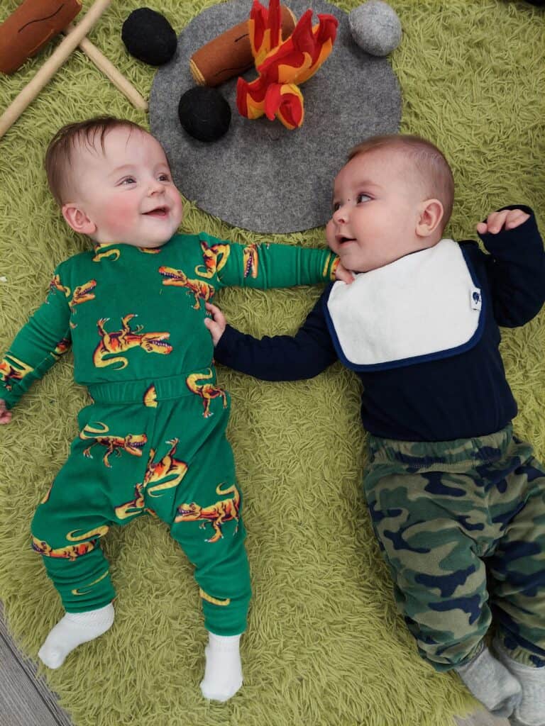 Two infants engaging in playtime on a green shaggy rug; one in a vibrant dinosaur jumpsuit and the other in a navy blue outfit with a bib, both looking at each other with bright, curious expressions.