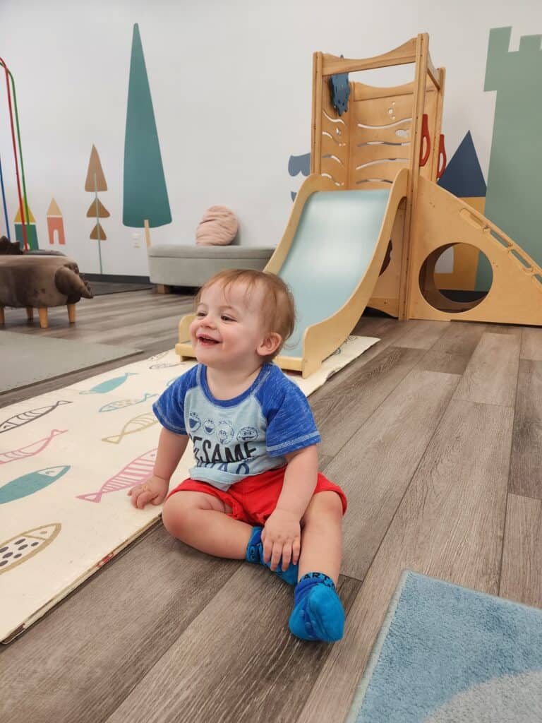 A joyful toddler wearing a blue 'Sesame' shirt and red shorts sits on a play mat at Bumble Brews in Cary, NC, surrounded by whimsical wooden play structures and soft-toned decor.