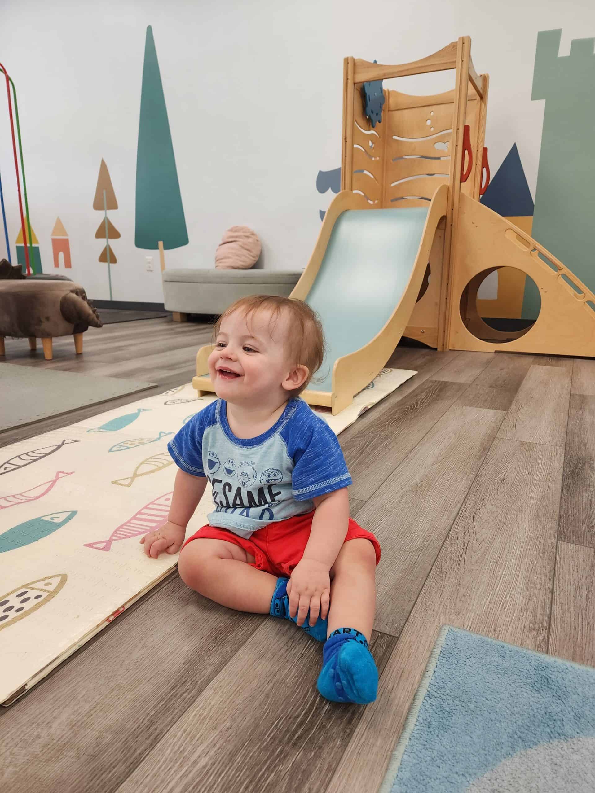A cheerful toddler sits on the floor wearing a 'Sesame' graphic tee and red shorts, laughing in a serene indoor playroom in Raleigh, NC, with minimalist wooden play structures and soft, muted tones in the background.