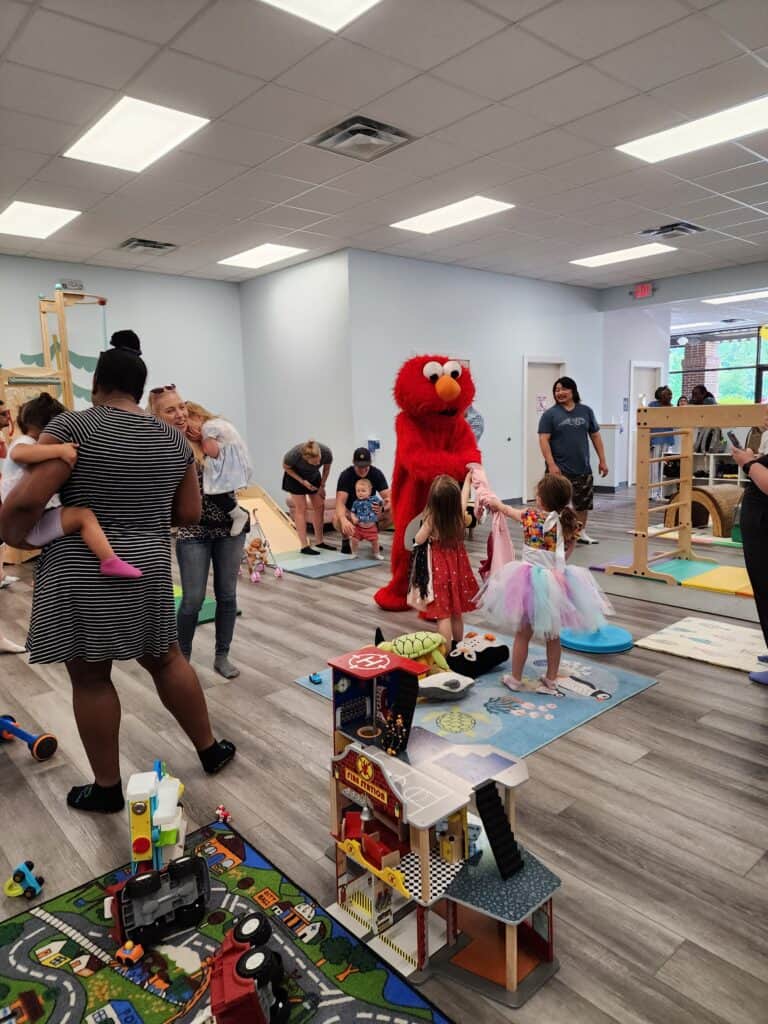 Bustling indoor play area with children and adults interacting, featuring a person in a bright red mascot costume, various toys, and a colorful road-themed rug, capturing a moment of joyful activity.
