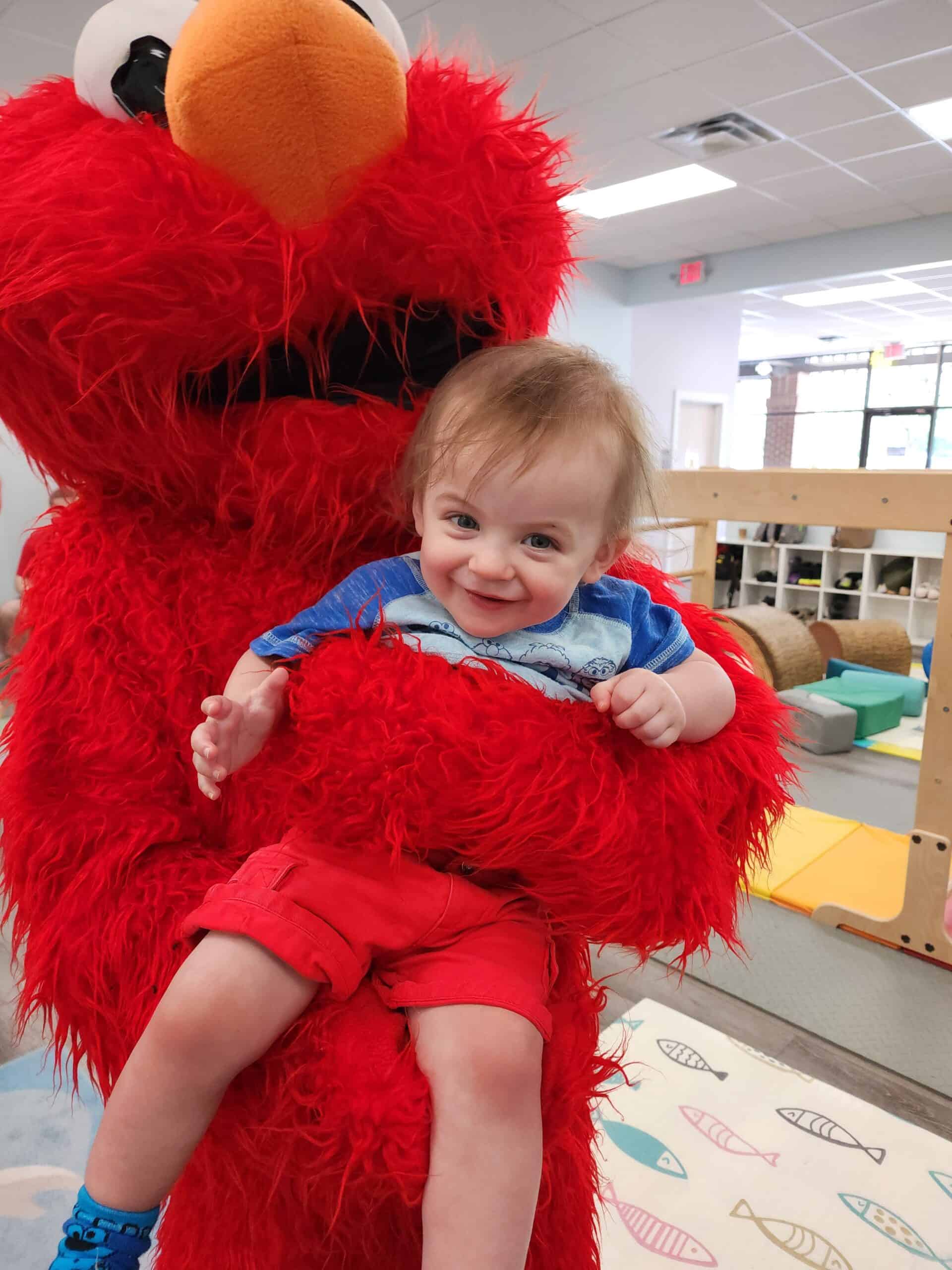 An elated toddler with a bright smile is held by a person in a fluffy, vibrant red costume at a children's indoor play area, conveying a moment of joy and playful interaction.