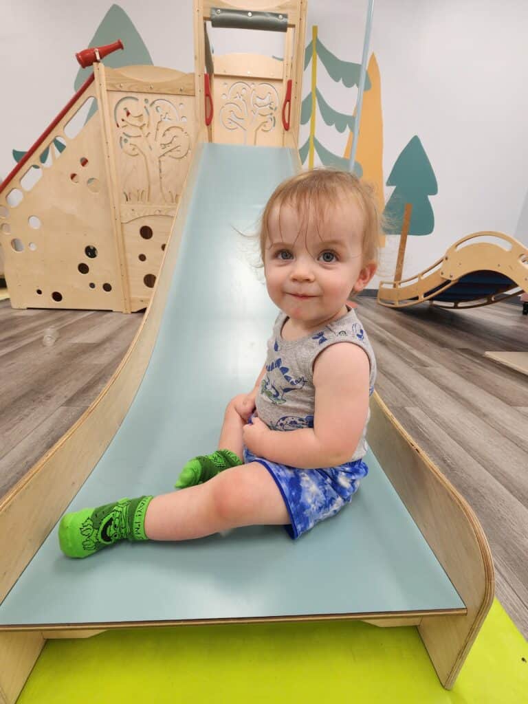 Adorable toddler with big blue eyes and a small smile, sitting on a light blue slide in a playful indoor setting with a wooden playset and tree-themed decorations in the background.