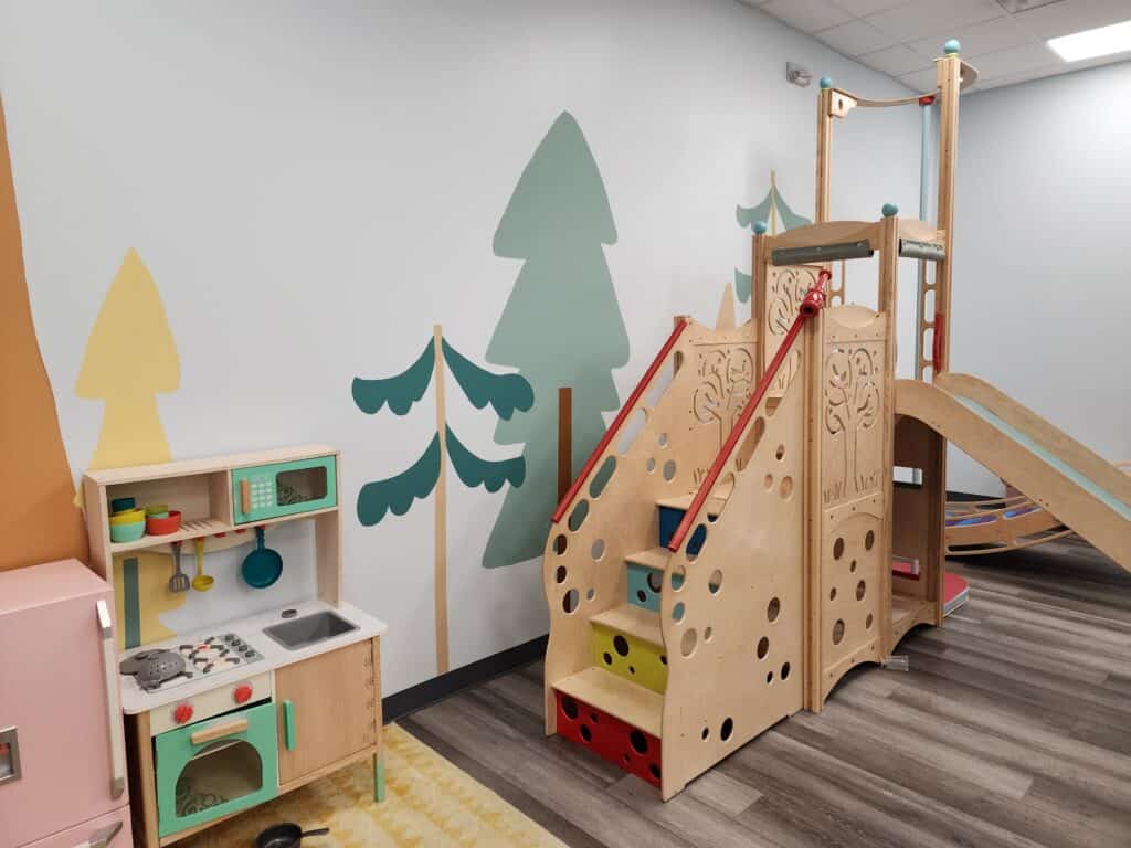 Bright and inviting indoor playground in Cary, NC, featuring a wooden play structure with a slide and climbing holes, complemented by colorful play kitchen accessories and whimsical tree murals.