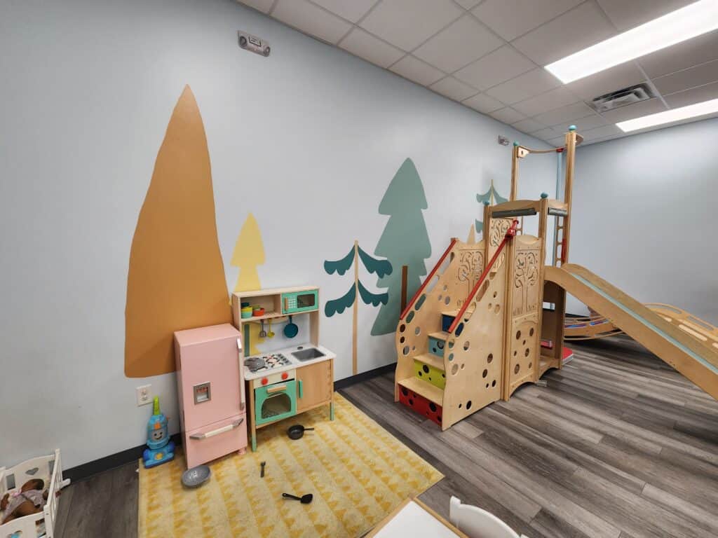 A modern indoor playroom designed for family fun in the Triangle area, featuring geometrically shaped wooden climbers, slides, and a play kitchen set against a minimalistic wall with abstract tree decals. The neutral tones and clean design invite imaginative play in a safe and welcoming environment