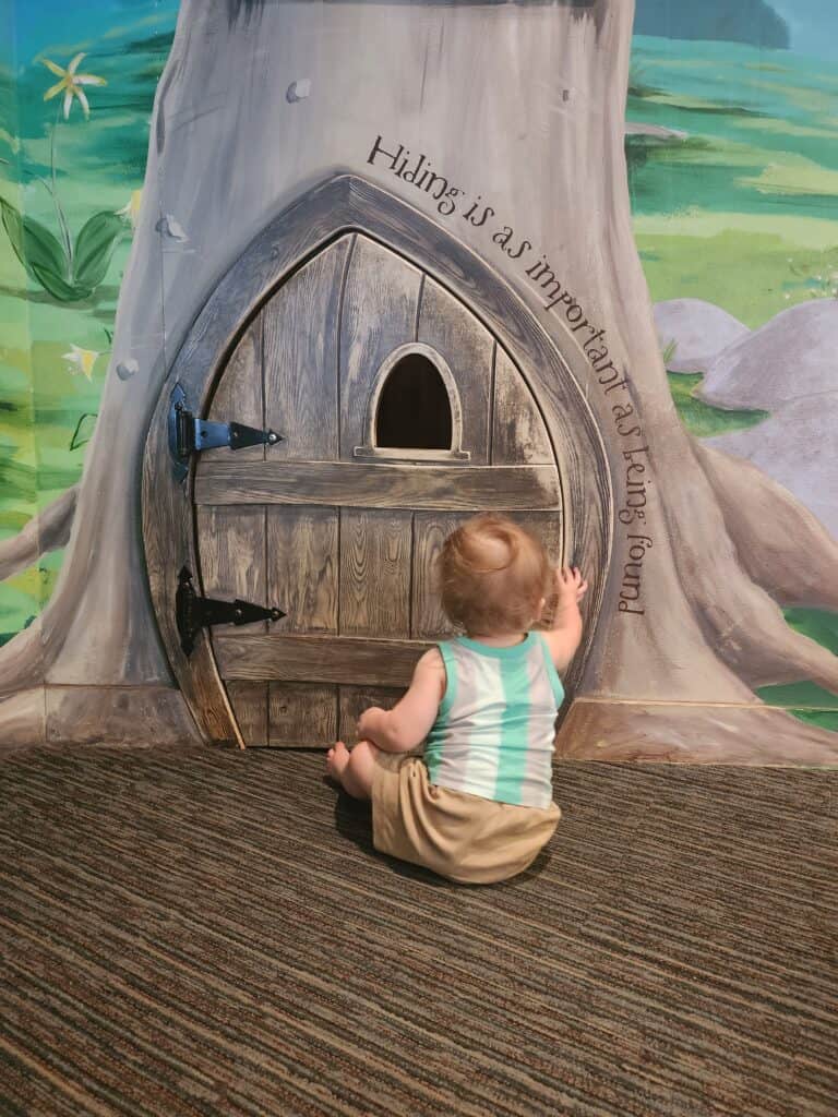 A curious toddler exploring Toddler's Hollow at Marbles Kids Museum in Raleigh NC, reaching for a whimsical door at the base of a painted tree, with the phrase 'Hiding is as important as being found' inscribed above