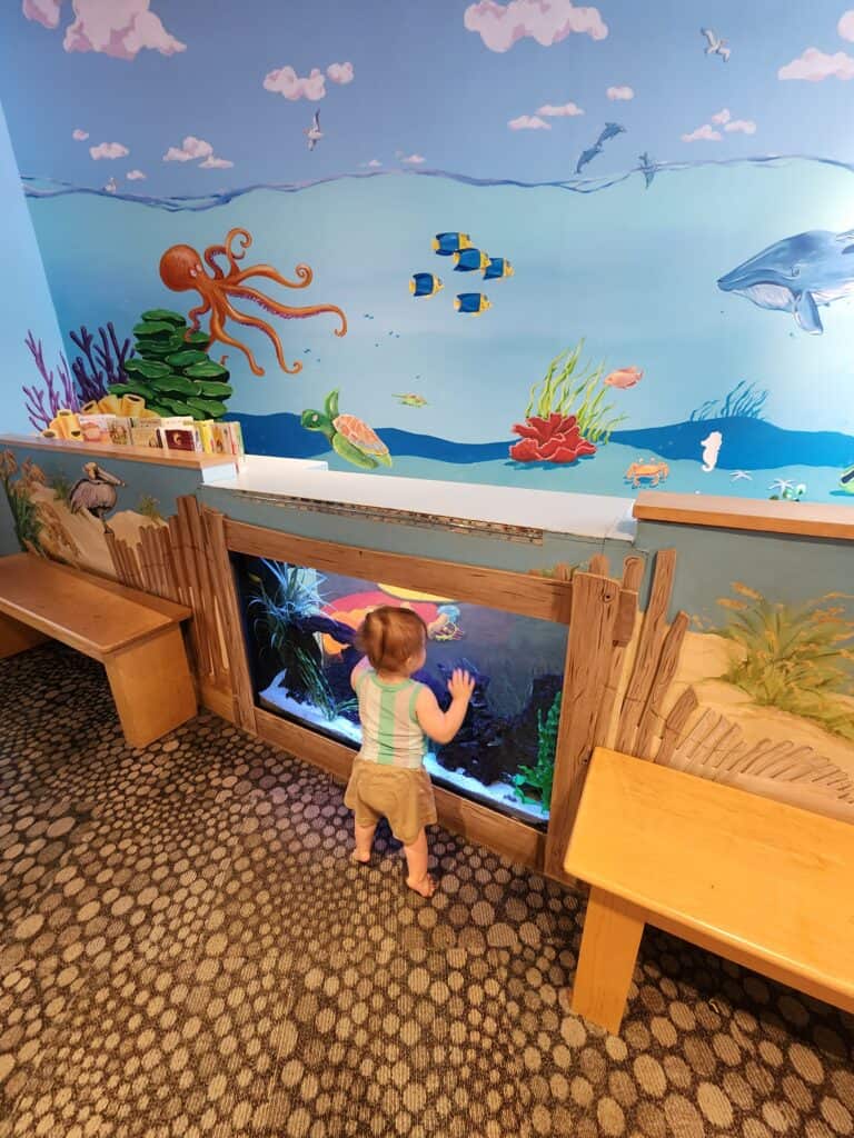 A young child is captivated by a colorful ocean-themed mural and an aquarium at a children's play area, illustrating one of the engaging things to do in Raleigh NC with kids. The mural depicts an underwater scene with a friendly octopus, fish, and a dolphin, creating a delightful and educational environment.