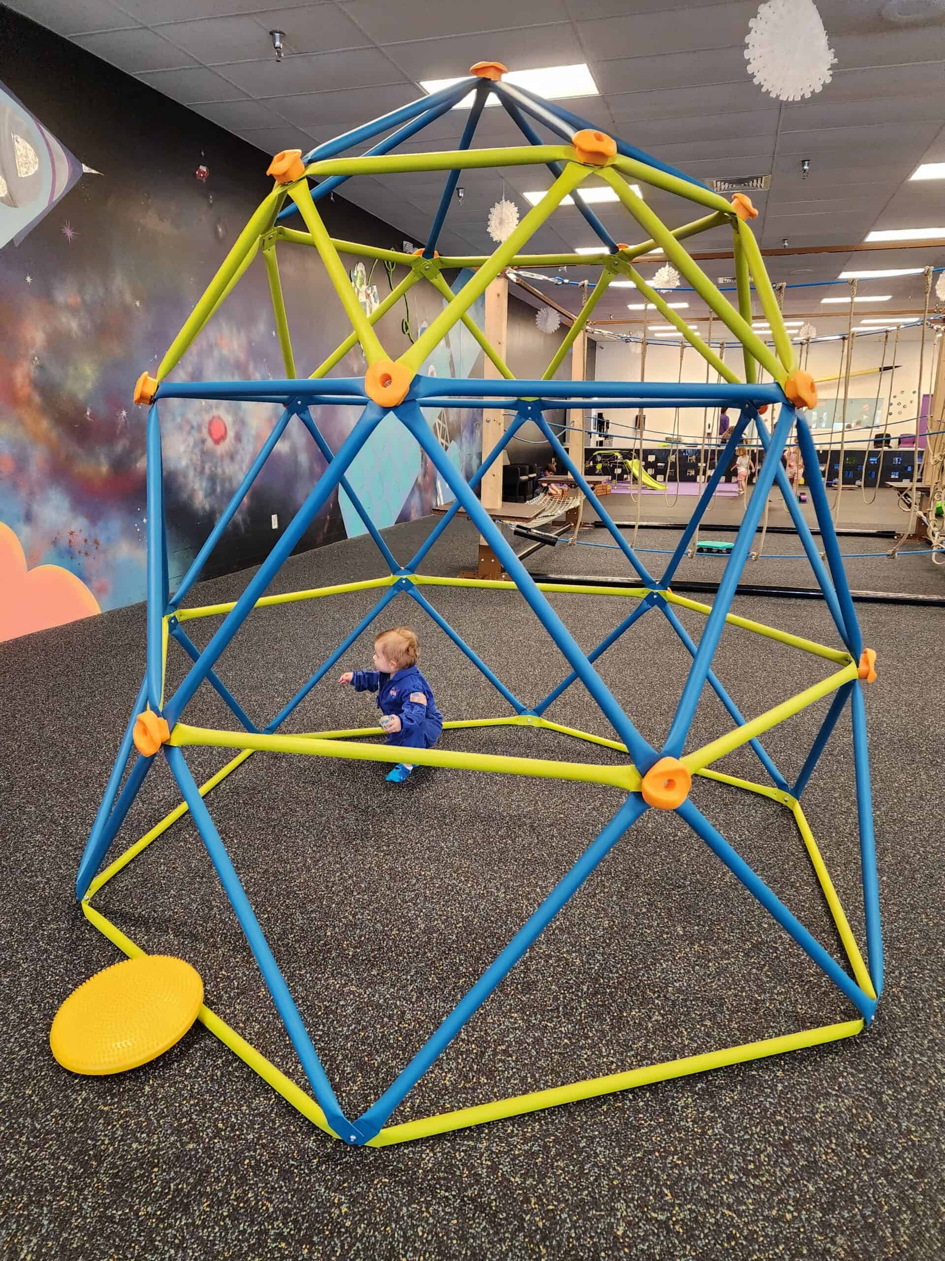 A small child in a blue outfit explores a vibrant, geometric climbing dome at Over The Moon Play Space in Cary, NC, with a colorful space-themed mural in the background.