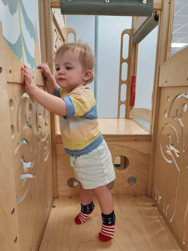 Inquisitive young child in a striped yellow and blue t-shirt and white shorts, exploring a wooden play structure with carved tree designs, wearing playful American flag-themed socks