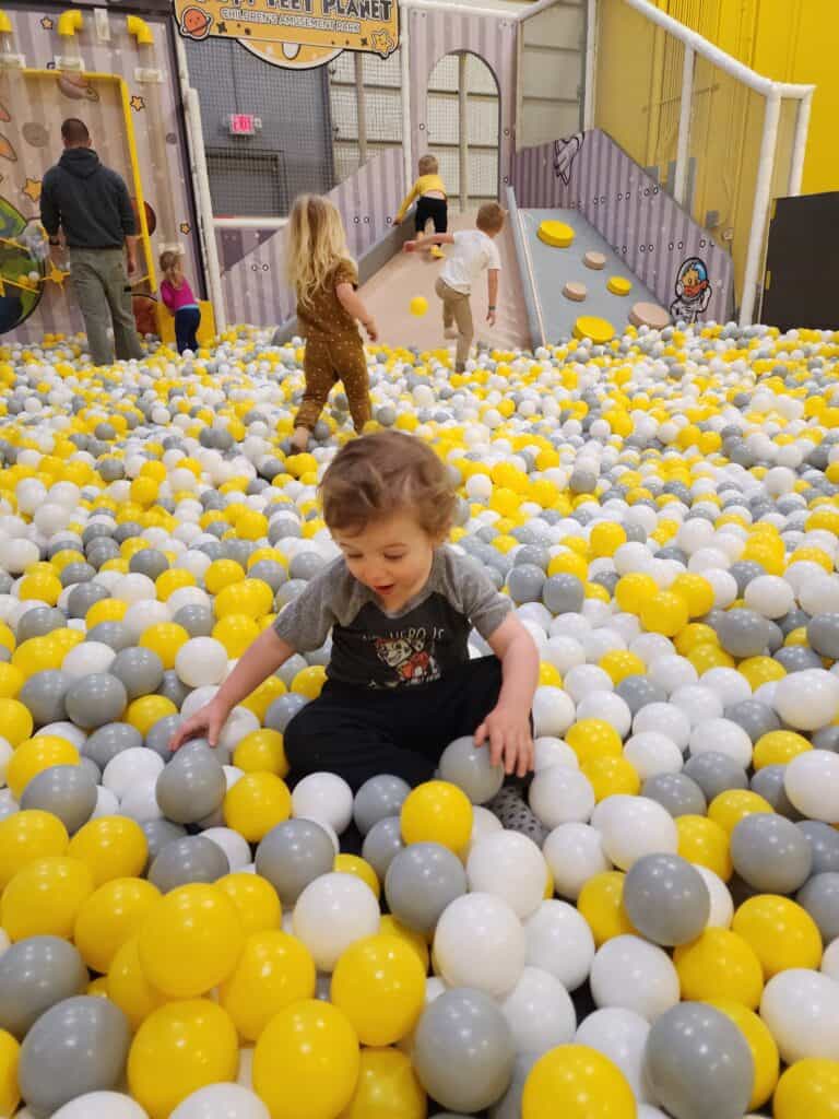 A young child with a playful expression sits among yellow and white balls at the Happy Feet Planet children’s amusement park in Apex, NC, with other children and adults in the background engaging with the play area
