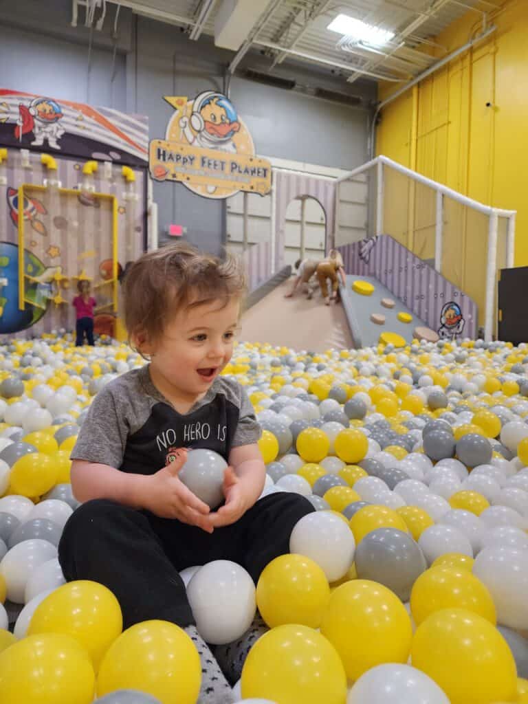 A delighted toddler sits among a sea of yellow and white balls in the ball pit at Happy Feet Planet, with the play center's cheerful signage in the background.