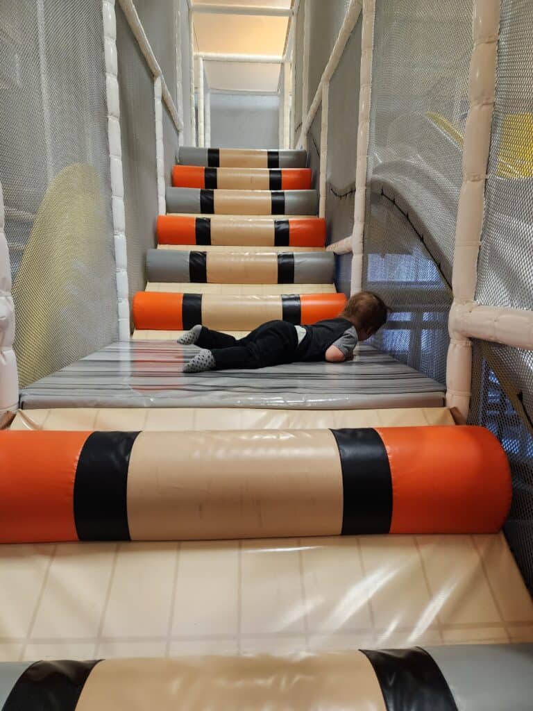 A young child lies playfully on soft, padded rollers within an obstacle course at an indoor playground near Raleigh, NC, enjoying a fun and safe play environment.