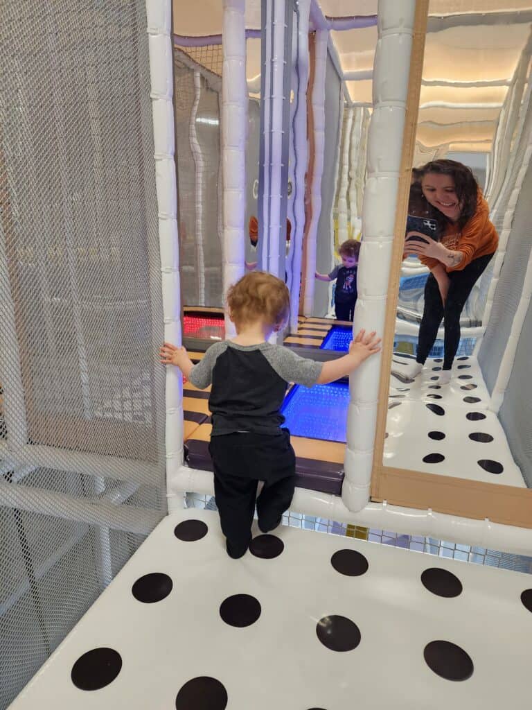 A toddler stands on a polka-dotted platform, reaching out to explore a vertical maze of light-up tubes, while a smiling adult assists another child in the background at an indoor play area in Raleigh, NC