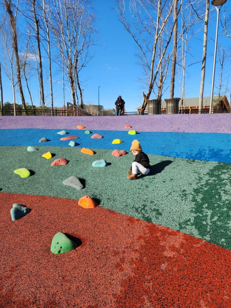 A child interacts with colorful climbing holds on a vibrant artificial rock-climbing mound at a playground, illustrating an engaging and tactile experience. The rubber safety surfacing transitions from blue to red, under the watchful eye of an adult, against a backdrop of tall leafless trees and a clear blue sky.