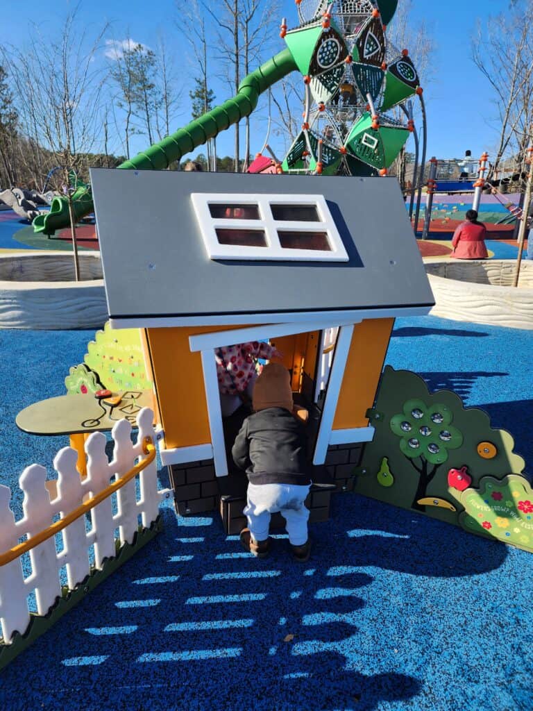 A child is seen entering a playhouse at a playground in Apex, NC, highlighting the park's interactive and child-friendly design. The playhouse is part of a larger play area that features a green tube slide and climbing structures in the background, all set on a bright blue safety surface under a clear blue sky.
