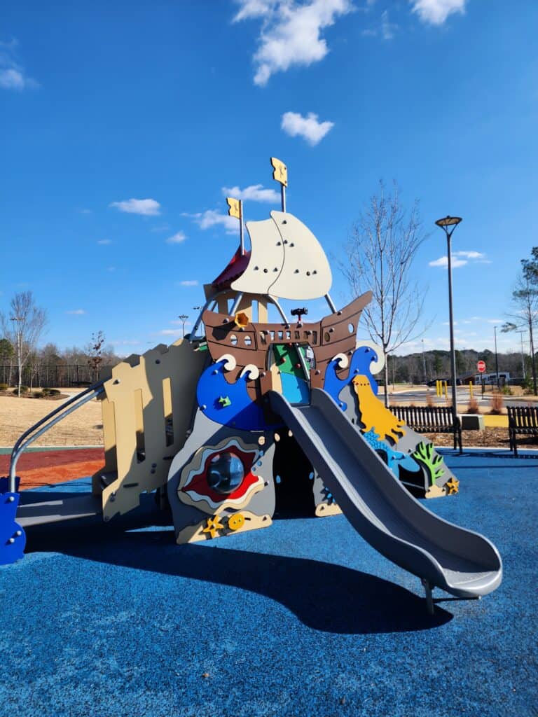 A whimsical pirate ship playground structure in Apex, NC, with playful sea creature designs and a gray slide, set against a bright blue rubber surface. The ship sports white sails with yellow flags, encouraging imaginative play under a sunny sky with sparse clouds