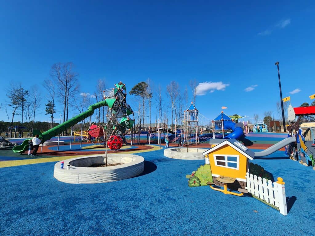 A wide-angle view of a modern and colorful playground with a variety of equipment including a green tube slide, climbing nets, and a whimsical playhouse with a white picket fence. The playground's surface is a mosaic of bright blue rubber, and the setting is a sunny day with clear skies, inviting active play in a safe and imaginative environment.