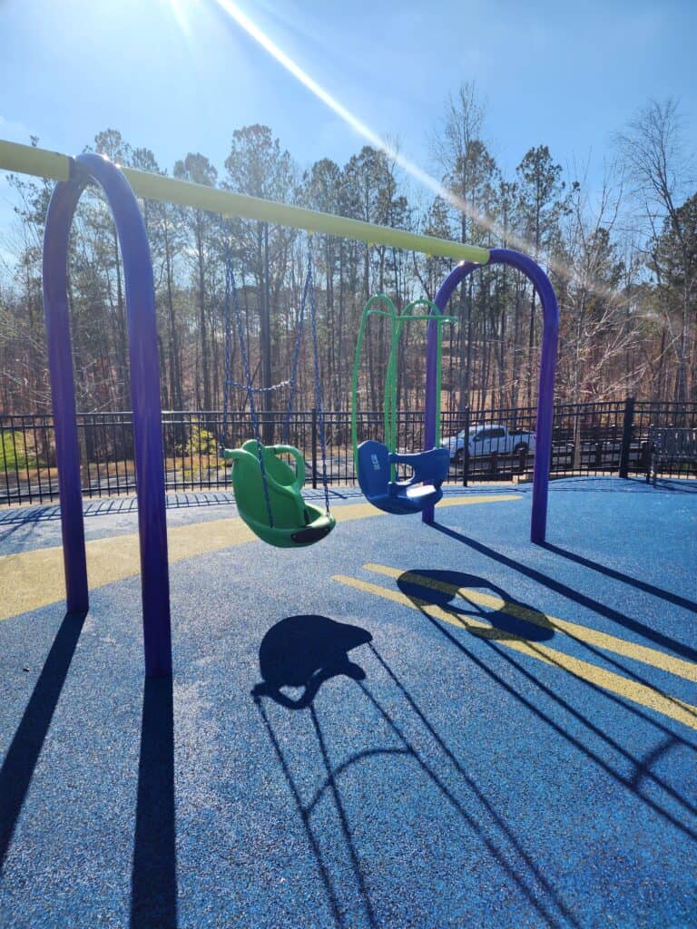 A peaceful playground scene featuring an accessible swing set with a green standard swing and a blue ADA swing, casting long shadows on the bright blue safety surface. The sun's rays add a warm touch to the tranquil outdoor space, surrounded by a fence and bare trees, inviting inclusive play on a sunny day.