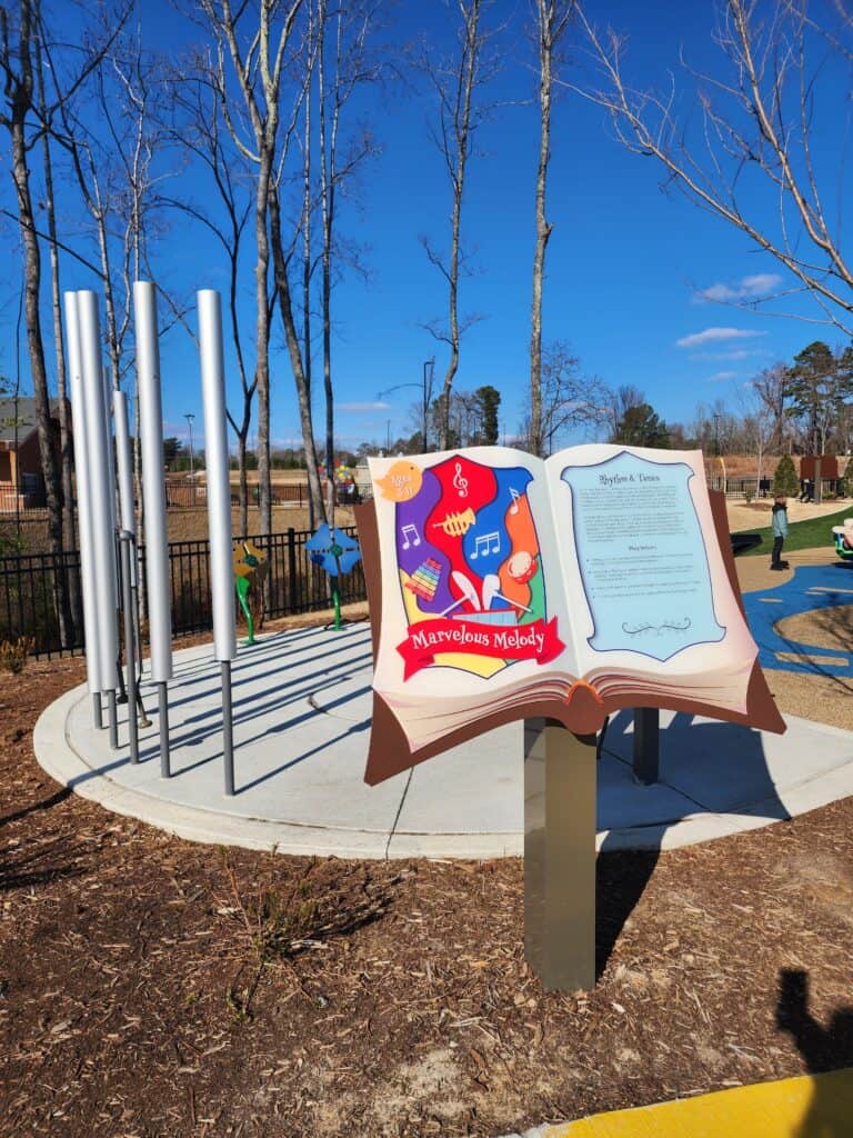 An interactive music-themed area at a Raleigh area playground, featuring a 'Marvelous Melody' installation with colorful illustrations and a series of vertical chimes. An open book-shaped sign provides educational content, inviting children to create music against the backdrop of a clear blue sky and leafless trees