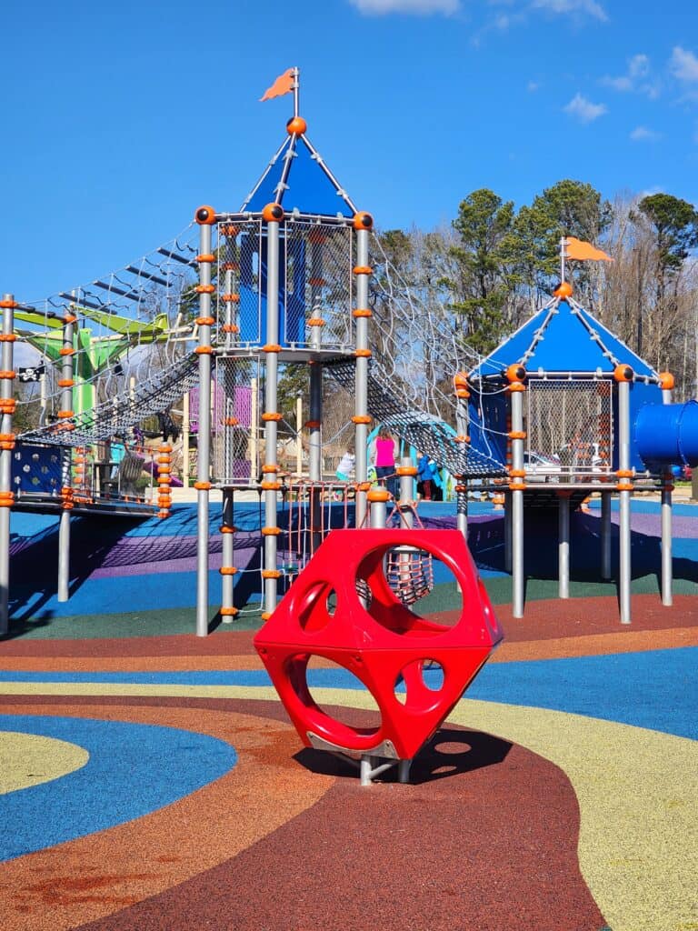 Colorful playground equipment at Pleasant Park in Apex, NC, with a focus on a red spherical climbing frame in the foreground. The background shows a series of interconnected play structures with blue roofs, climbing nets, and orange accents, all under a bright blue sky