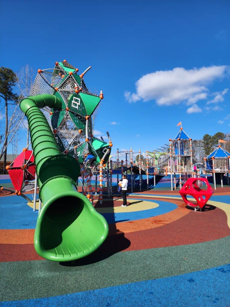A captivating playground near Raleigh, North Carolina, with a prominent green tube slide and intricate climbing structures. The play area is dotted with vibrant red, blue, and yellow safety surfacing, under a bright blue sky, inviting children for outdoor fun and adventure
