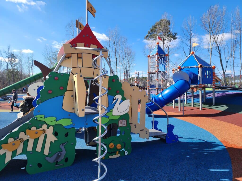 A playful castle-themed play structure with a spiral slide and various climbing elements at a sunny playground. The design includes sea creature motifs and a faux rock climbing wall, set against a vibrant blue safety surface, encouraging imaginative play in an outdoor setting with a bright blue sky