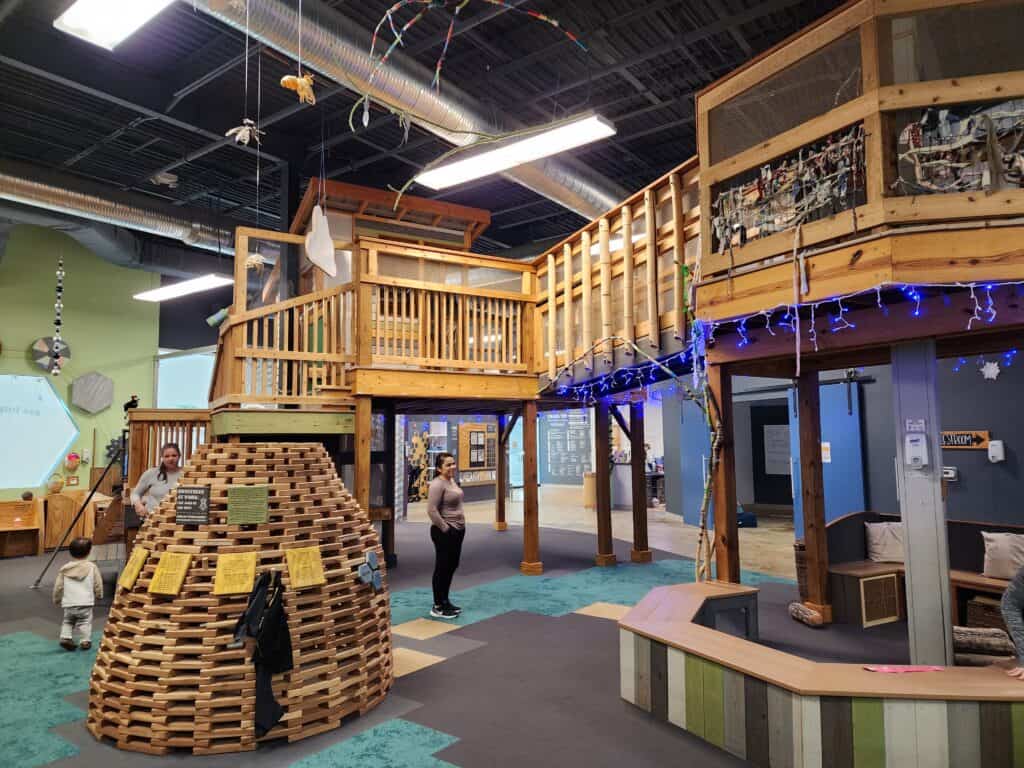 A dynamic Chapel Hill play place for kids featuring a wooden beehive climbing structure, a two-story playhouse, and interactive stations, all under a ceiling adorned with playful mobiles and soft glowing lights