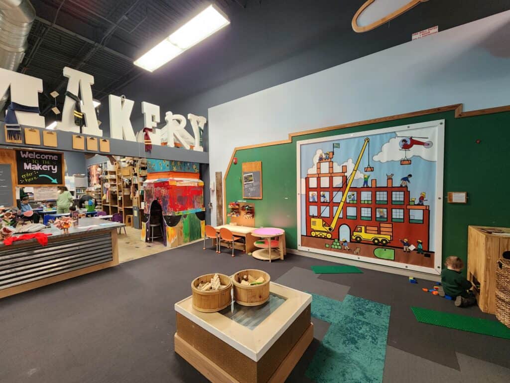 Inside the Kidzu Children's Museum in Chapel Hill, NC, the 'Makery' space buzzes with creativity, featuring a colorful crafting area, interactive wall puzzles, and a child engrossed in play with building blocks on a patterned floor mat