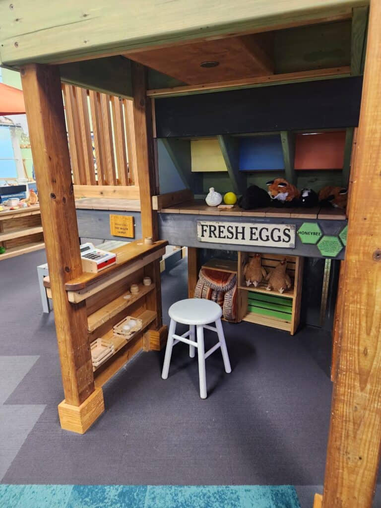 A cozy play market stall with a 'Fresh Eggs' sign, featuring wooden shelves stocked with play food and farm goods, a white stool, and educational materials, part of a child-friendly exhibit designed to simulate a farm-to-table experience