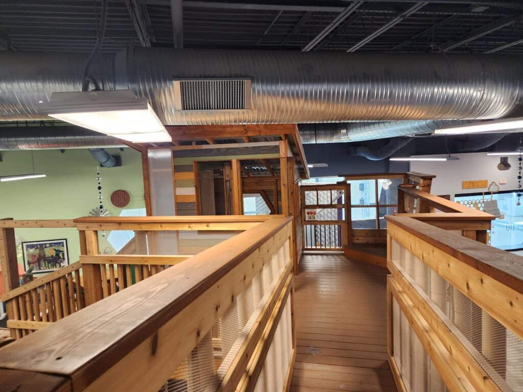 Elevated wooden walkways inside a children's museum in Chapel Hill, providing a bird's-eye view of various interactive exhibits below, encouraging exploration and physical play in a safe, indoor environment