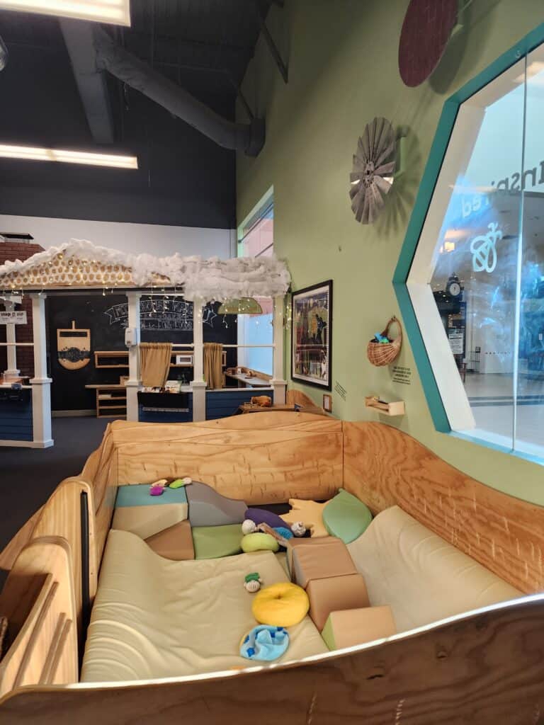 An imaginative indoor play area in Chapel Hill, designed with a soft, cushioned pit filled with colorful shapes and textures, adjacent to an interactive exhibit, encouraging playful exploration and physical activity for children.