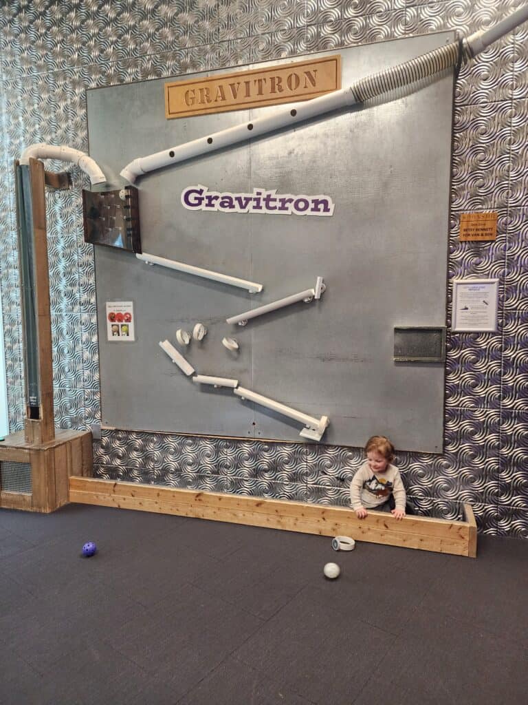 A child experiments with the laws of physics at the 'Gravitron' exhibit in an indoor playground in the Triangle area, showcasing a fascinating ball track on a metal wall, designed to teach gravity and motion in an interactive and fun way.