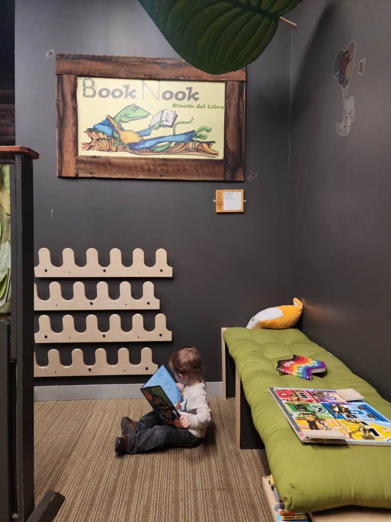 A child sits engrossed in reading at the 'Book Nook' of the Kidzu Children's Museum in Chapel Hill, NC, with whimsical wall art and a cozy green reading bench, creating a peaceful corner for young readers to dive into stories