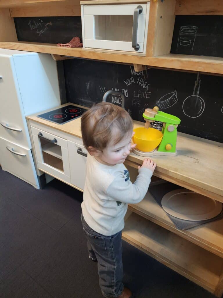 A young child plays at a miniature kitchen station with a chalkboard backsplash, creatively engaging with a toy blender and yellow bowl, fostering imaginative play in a child-friendly space