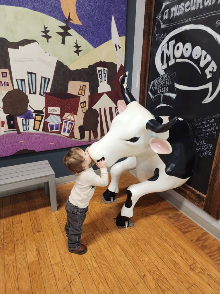 A curious child interacts with a lifelike cow sculpture by giving it a kiss, at a museum exhibit in Chapel Hill, under a mural depicting a whimsical town scene, evoking a sense of discovery and fun in a hands-on learning environment.